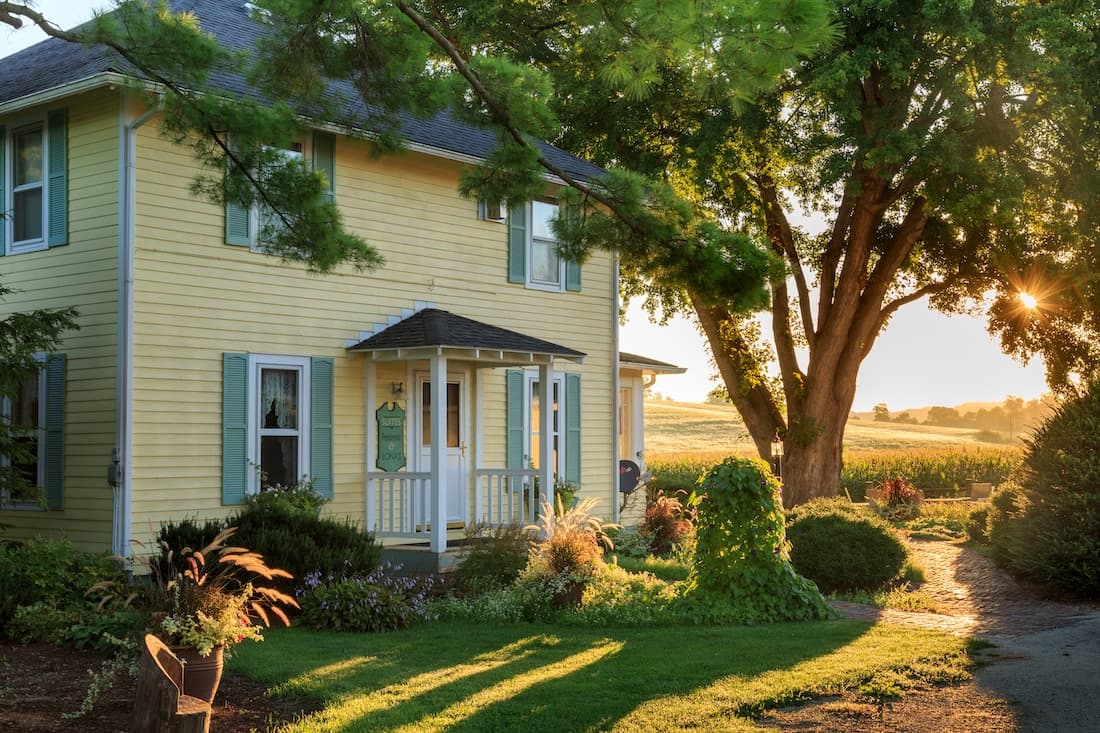 A sunlit yellow house surrounded by lush greenery and gardens.