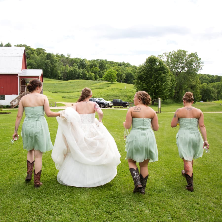 A bride dressed in a white wedding dress, and three bridesmaids dressed in green dresses and cowboy boots, walking towards a barn