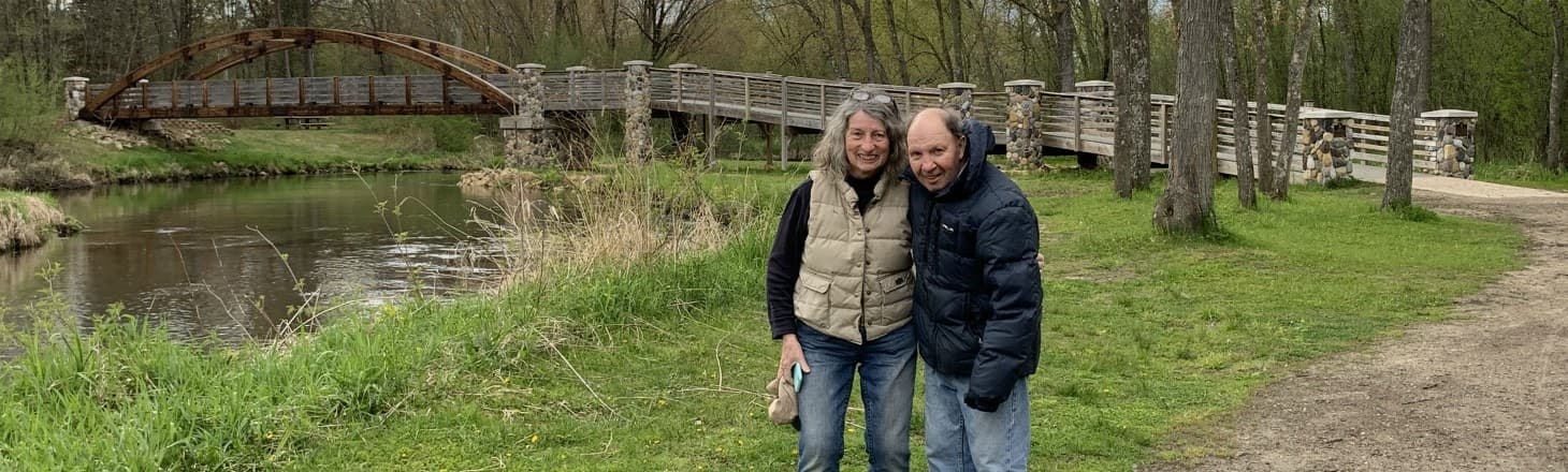A couple stands smiling on a path near a bridge and river surrounded by greenery.