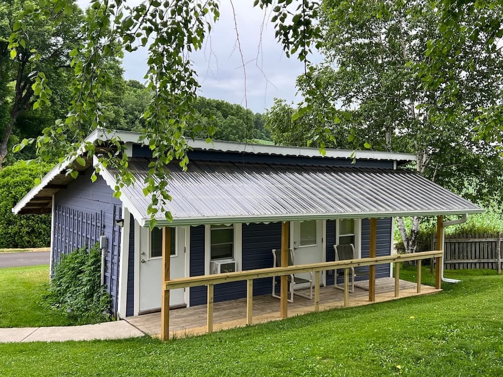 A small, blue cabin with a metal roof and a front porch surrounded by greenery.