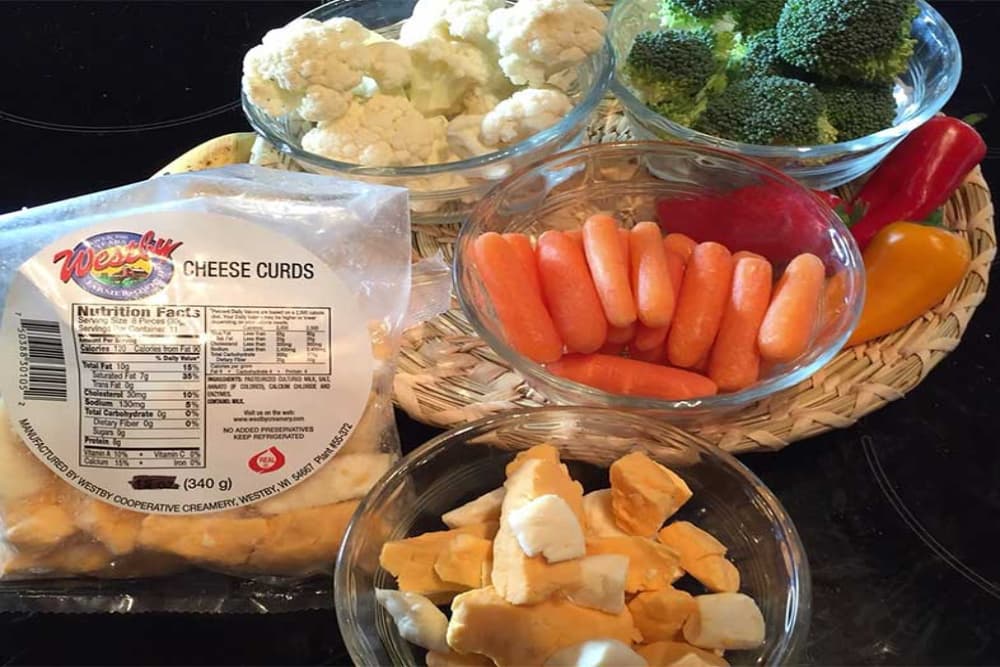 A wicker basket with a variety of fresh vegetables, and a package of white and orange Wisconsin cheese curds