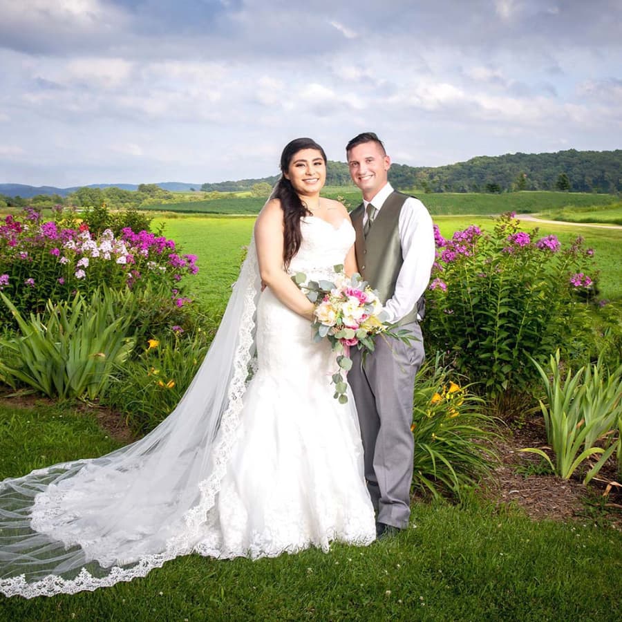 A smiling groom posing for pictures outside with beautiful white and pink flowers in the garden on either side of them.