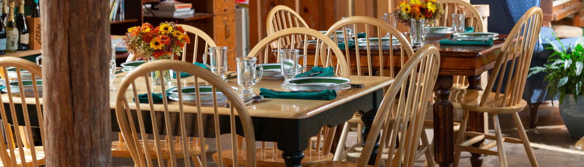 A room with 2 wood tables and chairs set with china, green napkins, and colorful flowers