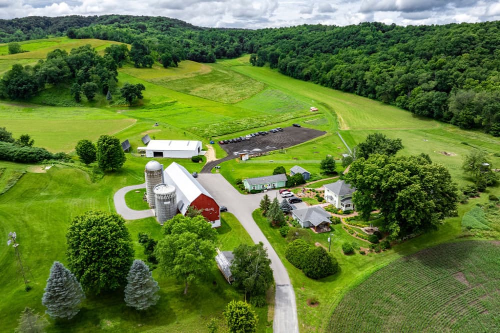 Aerial view of farm with red barn, silos, other outbuildings, green grass and trees with meandering lane.