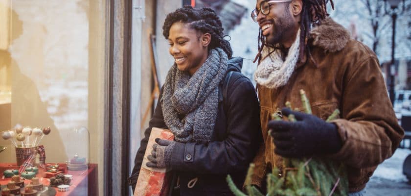 A smiling couple stands outside a shop looking at holiday displays, with one holding a wrapped gift and the other carrying greenery.