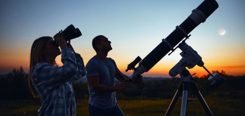 Two people observe the night sky, one using binoculars and the other looking through a telescope, against a sunset backdrop.