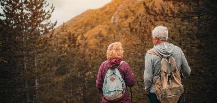 Two hikers, an older woman with a light pink backpack and an older man in a gray sweater, admire a mountain view surrounded by autumn trees.