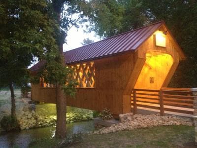 A wooden covered bridge with a red metal roof illuminated by lights crosses over a small stream surrounded by trees.