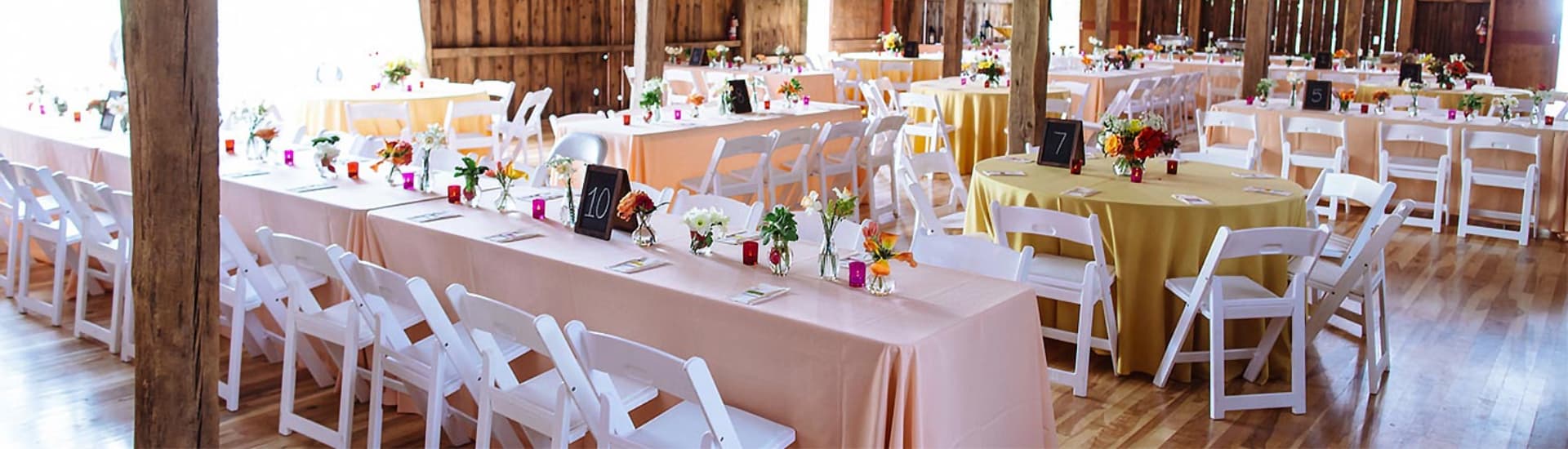 A beautifully arranged barn setting with white chairs and tables draped in pastel linens, adorned with colorful flower centerpieces.