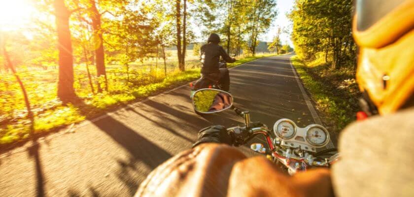 two motorcycle riders riding along a rural, tree-lined road with morning sun streaming through the trees.