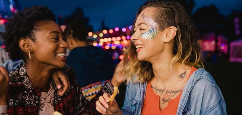 two friends laughing and enjoying food at an outdoor festival at night, with colorful lights in the background