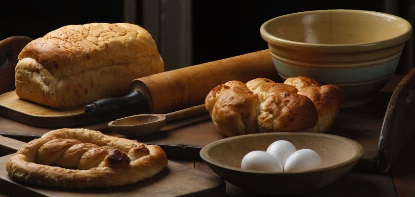 Fresh baked Amish breads, rolls, eggs, and a rolling pin arranged on a rustic wooden table.