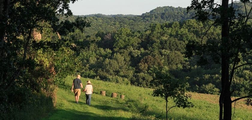 Two guests walk along a grassy trail at Justin Trails with wooded Driftless Region hills in the distance.